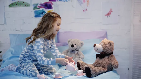 Side view of smiling preteen kid pouring tea near soft toys on bed — стоковое фото