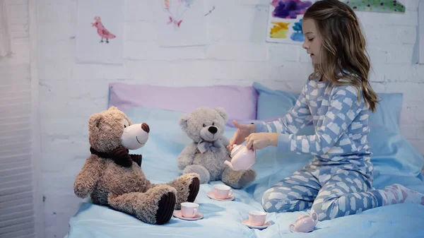Side view of kid in pajama pouring tea near teddy bears on bed - foto de stock