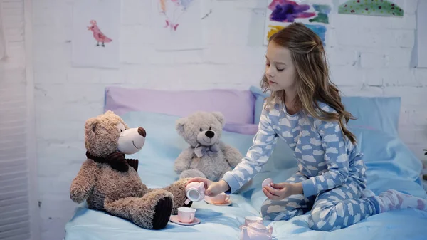 Child in pajama playing with tea cups and teddy bears on bed in evening — Photo de stock