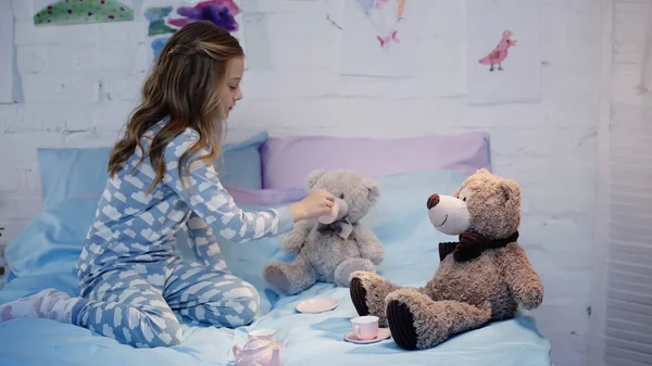 Side view of preteen kid in pajama holding cup near soft toy on bed — Fotografia de Stock