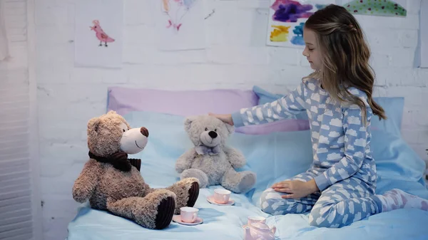 Side view of child in pajama touching sot toy near tea cups on bed at home — Stock Photo