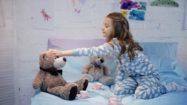 Side view of preteen kid touching soft toy near tea cups on bed in evening — стоковое фото