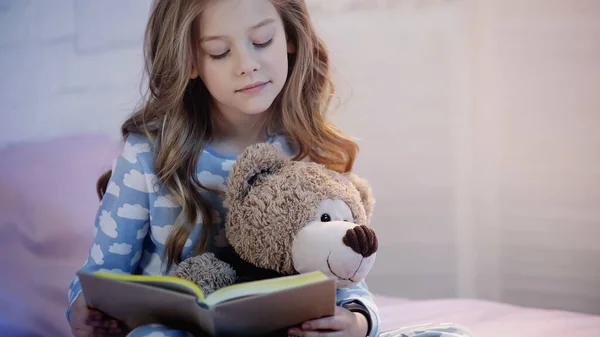 Preteen kid holding soft toy while reading book in bedroom — Stockfoto