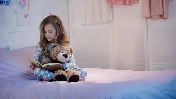 Preteen child in pajama reading book and hugging teddy bear in bedroom in evening — стоковое фото