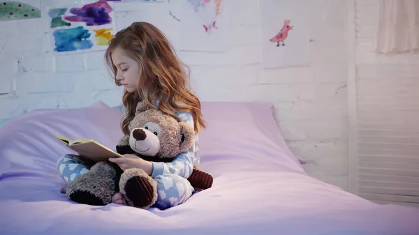 Side view of kid in pajama reading book and holding soft toy on bed at home — Stockfoto