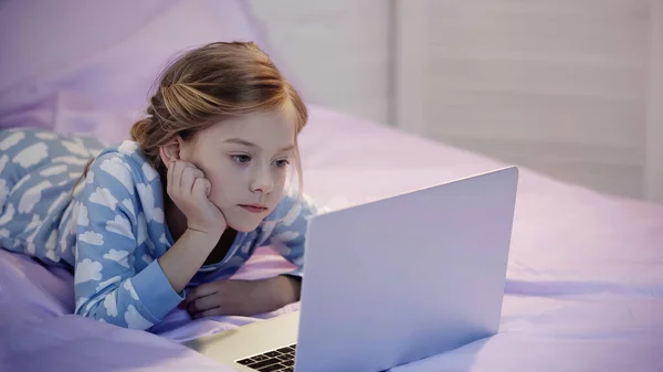 Preteen girl in pajama looking at laptop on bed in evening - foto de stock