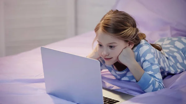 Smiling kid in pajama looking at laptop on bed in evening — Foto stock