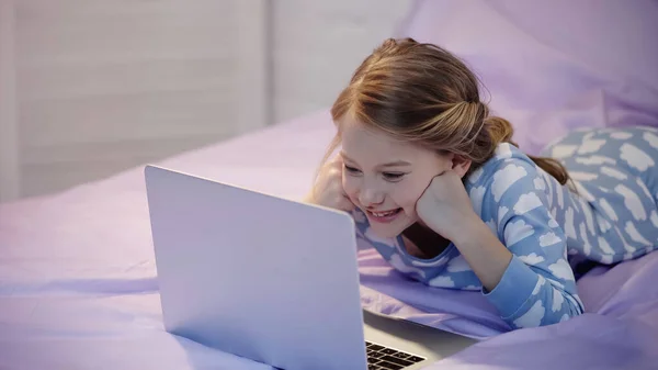 Glückliches Kind im Pyjama schaut abends auf Laptop im Schlafzimmer — Stockfoto