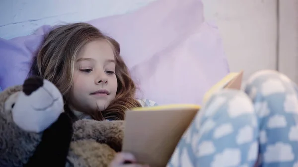 Preteen kid reading book while lying near teddy bear on bed — Stock Photo