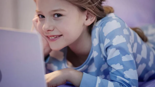 Smiling preteen kid in pajama looking at camera near blurred laptop in bedroom — Stock Photo