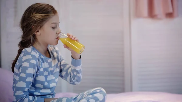 Preteen girl in pajama drinking orange juice on blurred bed in evening - foto de stock