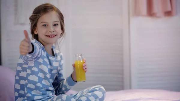 Smiling kid in pajama showing thumb up and holding orange juice on bed — Foto stock
