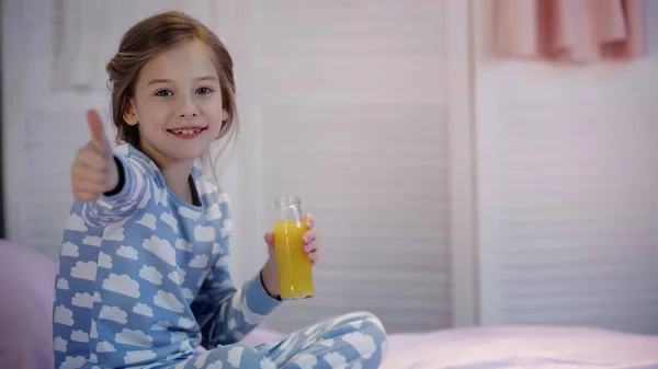 Smiling kid in pajama showing thumb up and holding orange juice on bed — Stock Photo