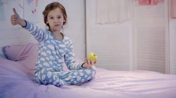 Kid in pajama showing like and holding apple on bed in evening — Stock Photo