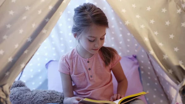 Preteen child in t-shirt reading book in wigwam in evening — стоковое фото