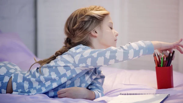 Side view of preteen child taking color pencil near sketchbook on bed — Fotografia de Stock