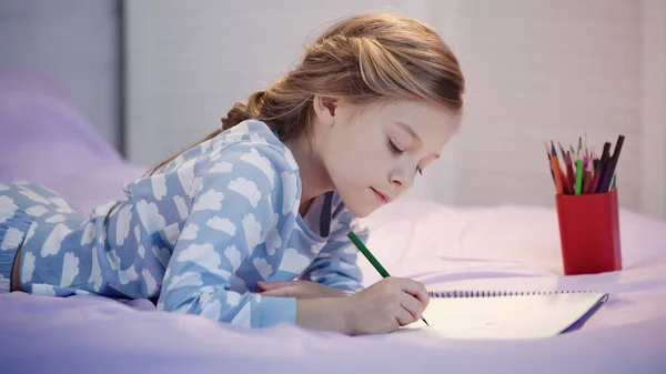 Preteen child drawing on sketchbook near color pencils on bed in evening — Photo de stock