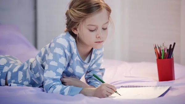 Preteen child in pajama drawing with color pencil on sketchbook on bed — Stockfoto