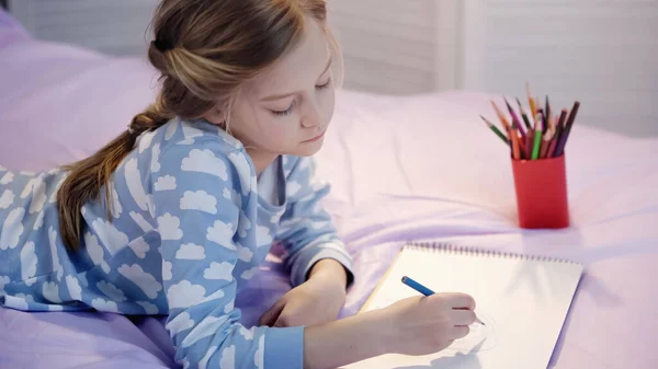Preteen girl drawing on sketchbook near blurred color pencils on bed — Fotografia de Stock