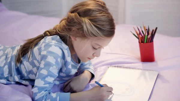 Preteen kid drawing on sketchbook while lying on bed in evening - foto de stock