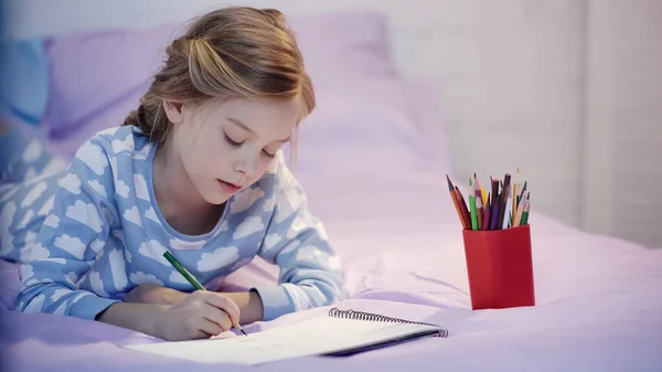 Kind im Pyjama zeichnet Skizzenbuch auf verschwommenem Bett am Abend — Stockfoto