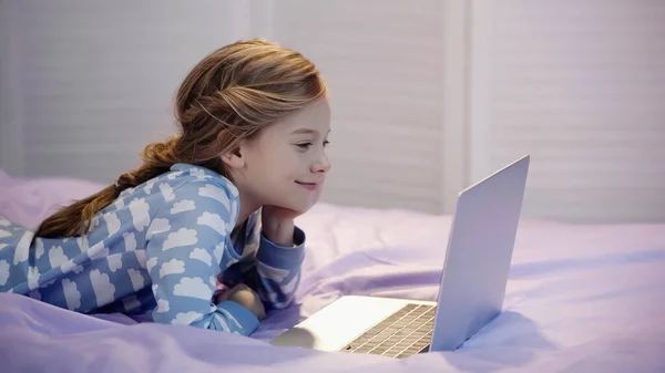 Smiling girl in pajama looking at laptop in bedroom in evening — Stock Photo