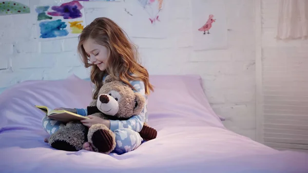 Happy preteen kid reading book and hugging soft toy on bed in bedroom — Foto stock