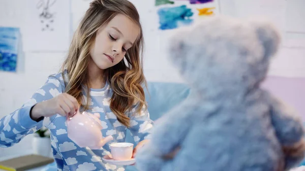 Preteen child in pajama pouring tea near blurred soft toy in bedroom - foto de stock