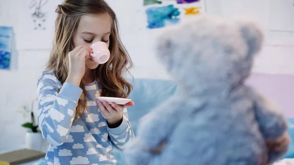 Child in pajama drinking tea near blurred teddy bear in bedroom — Fotografia de Stock