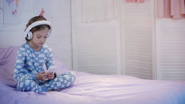 Preteen child in pajama using headphones and smartphone on bed in evening - foto de stock