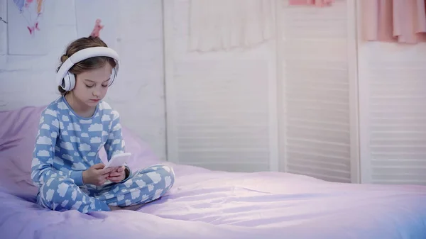 Preteen child in pajama using headphones and smartphone on bed in evening — Photo de stock