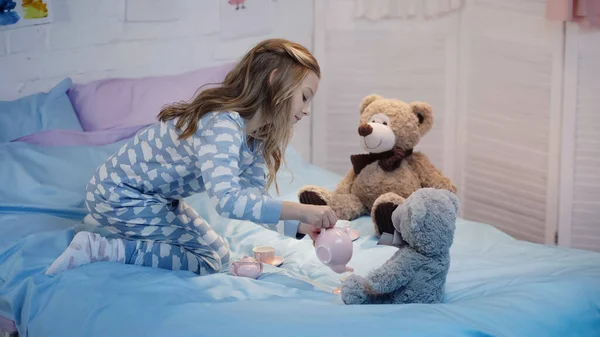 Side view of preteen child pouring tea while playing near teddy bears on bed — стоковое фото