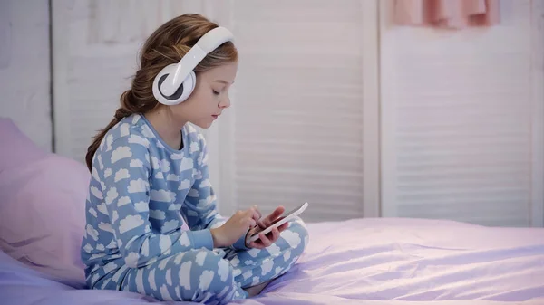 Side view of child in headphones using cellphone on bed - foto de stock