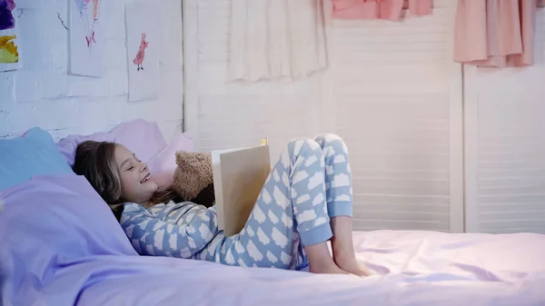 Preteen kid in pajama laughing while reading book near teddy bear on bed — Fotografia de Stock