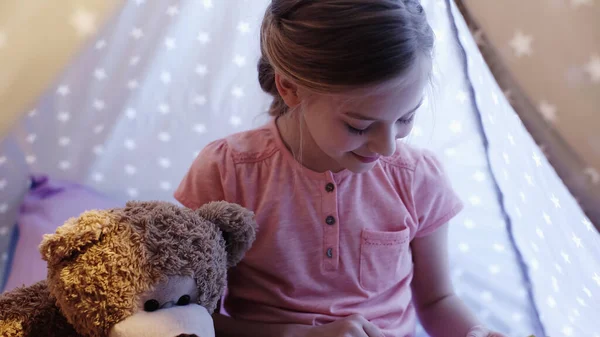 Smiling child looking away near soft toy in wigwam in evening — Stock Photo