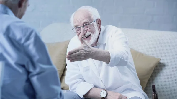 Старший бородатий чоловік з простягнутою рукою посміхається біля розмитого друга вдома — стокове фото