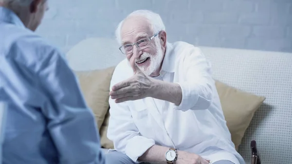 Senior bearded man with outstretched hand smiling near blurred friend at home — Stockfoto