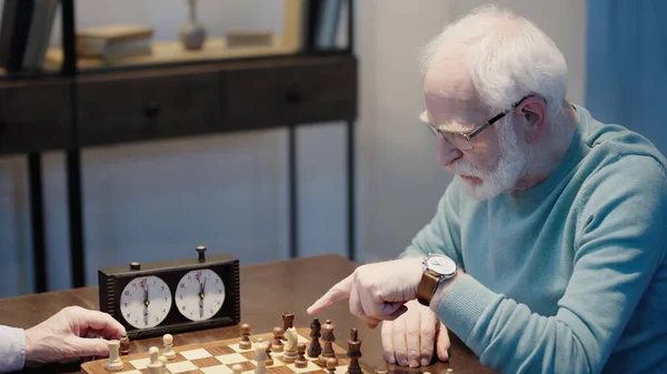 Elderly man pointing at chessboard near timer and friend — Stockfoto