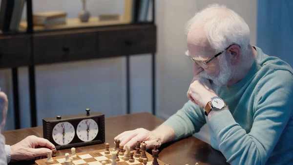 Thoughtful senior man moving chess figure while gaming with friend in living room — Stock Photo