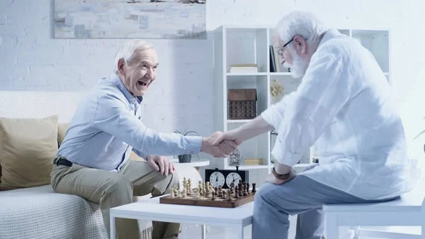 Excited man shaking hands with senior friend while playing chess in living room — Stockfoto