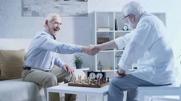 Happy senior man shaking hands with friend near chessboard in living room — Stockfoto