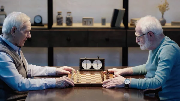 Side view of senior grey haired friends playing chess in living room - foto de stock