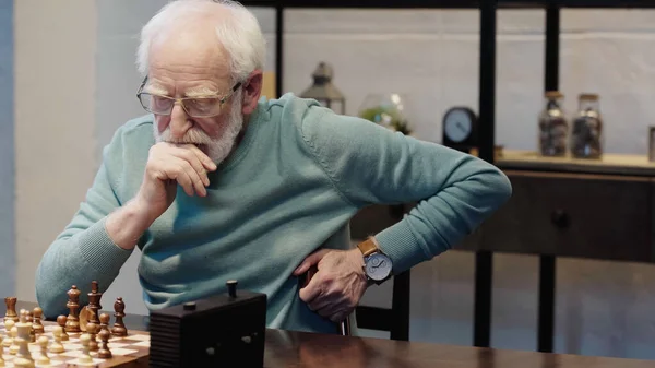 Senior man in eyeglasses thinking near chessboard at home — Stockfoto