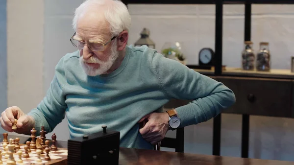 Senior bearded man holding chess figure while gaming in living room - foto de stock