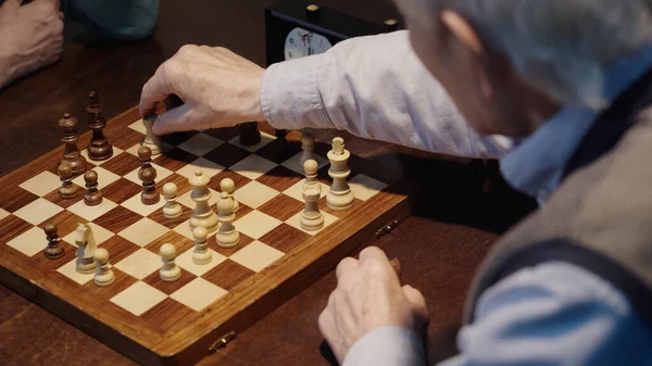 Cropped view of blurred senior man moving figure on chessboard while playing with friend - foto de stock