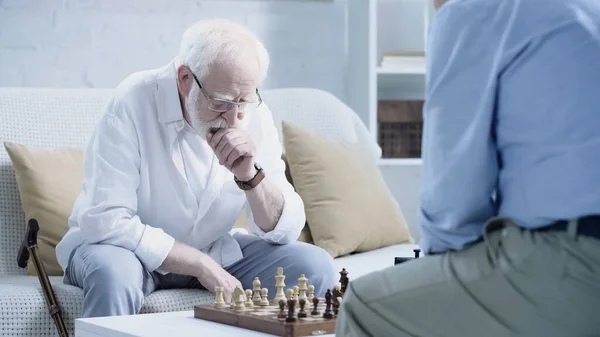 Elderly man in eyeglasses thinking near chessboard and blurred friend in living room — Fotografia de Stock