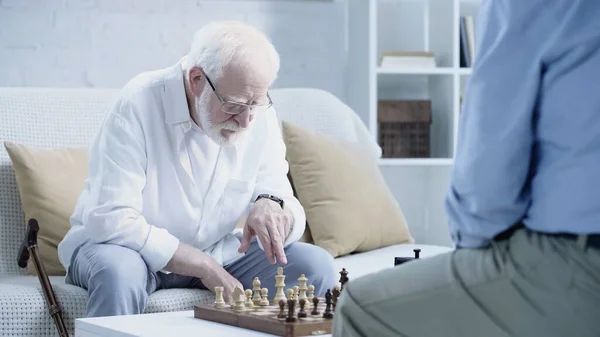 Senior man in eyeglasses pointing at chessboard near blurred friend — Stock Photo