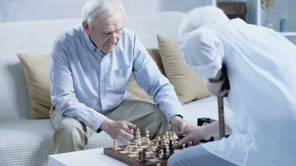 Senior man sitting on couch and playing chess with blurred friend - foto de stock