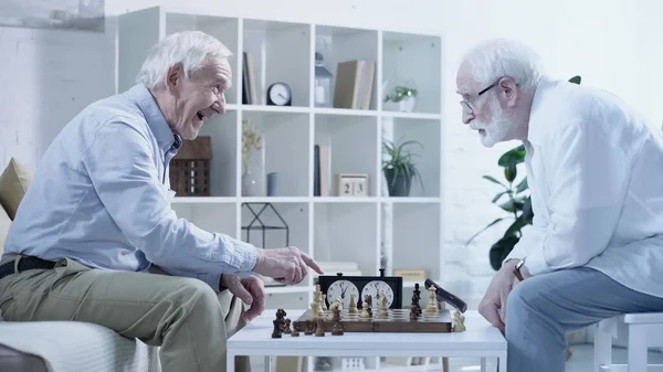 Side view of laughing man pointing at chessboard near senior friend — стоковое фото