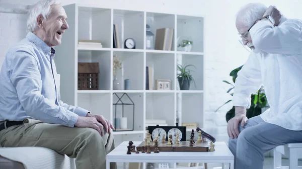 Side view of senior man laughing near chessboard and thoughtful friend — Stock Photo